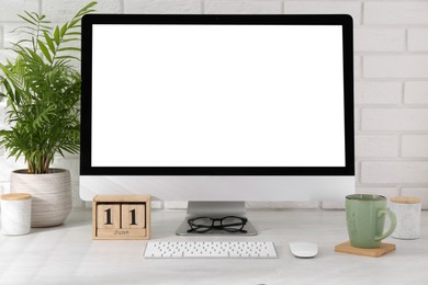 Office workplace with computer, glasses, cup, wooden block calendar and houseplant on light table near white brick wall