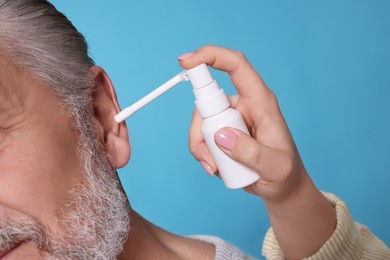 Young woman spraying medication into man's ear on light blue background, closeup