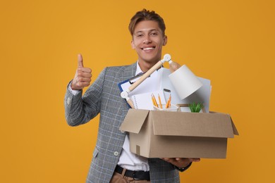 Photo of Happy unemployed young man with box of personal office belongings showing thumb up on orange background
