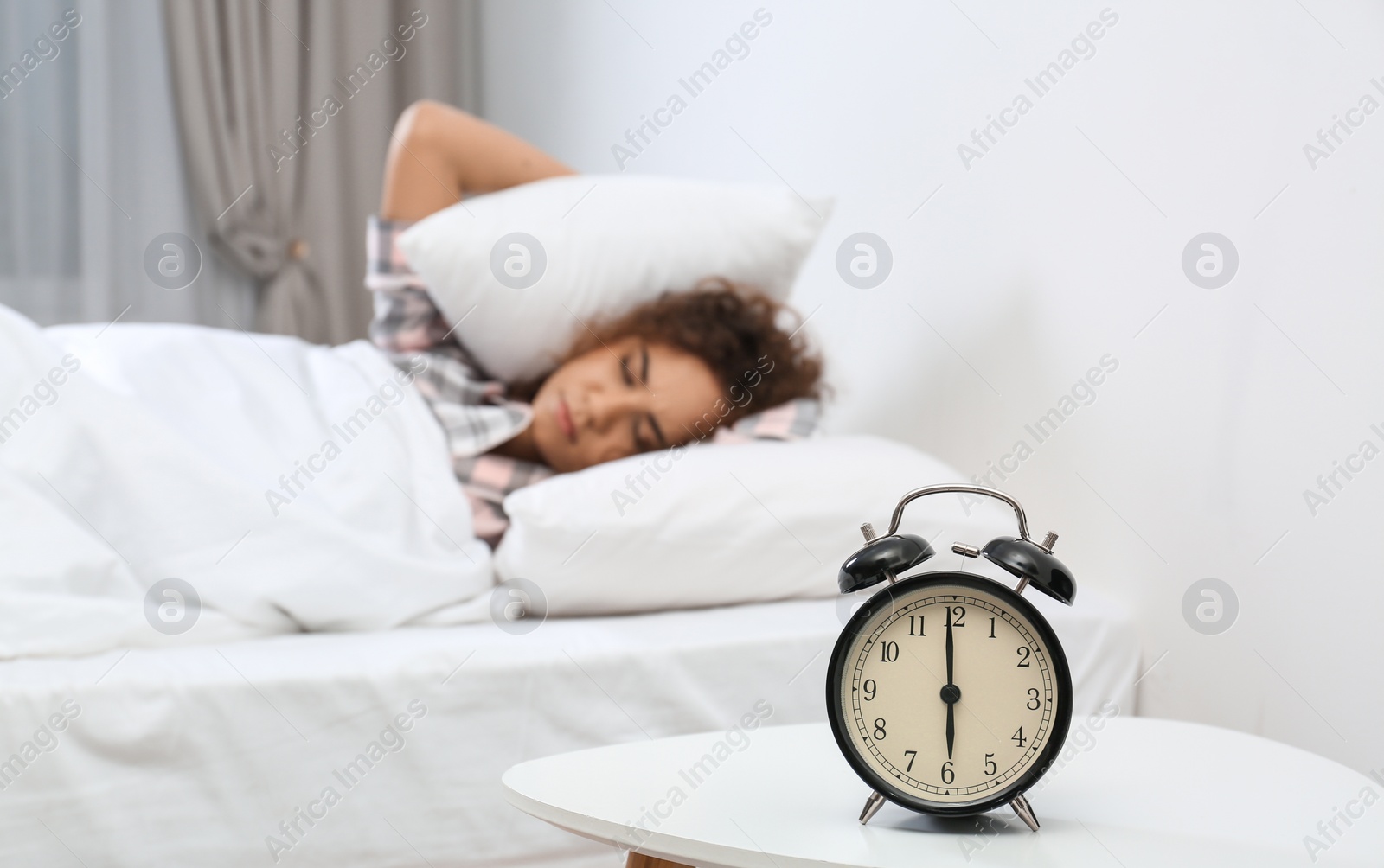 Photo of Young African-American woman covering ears with pillows while hiding from alarm clock at home. Bedtime