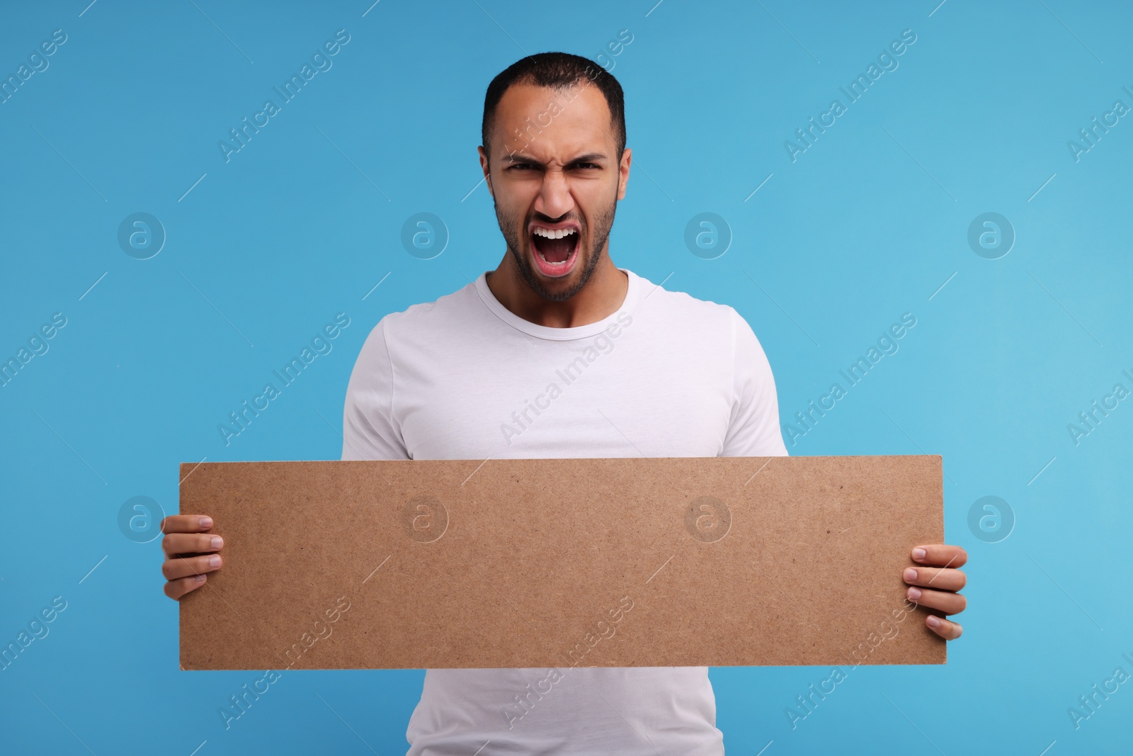 Photo of Angry man holding blank cardboard banner on light blue background, space for text