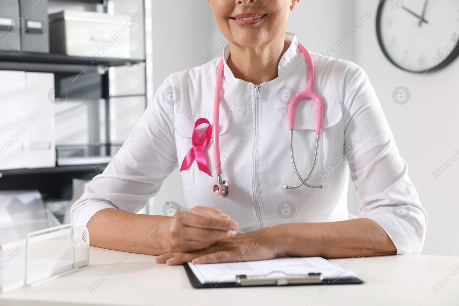 Photo of Doctor with stethoscope and pink ribbon at white desk indoors, closeup. Breast cancer awareness