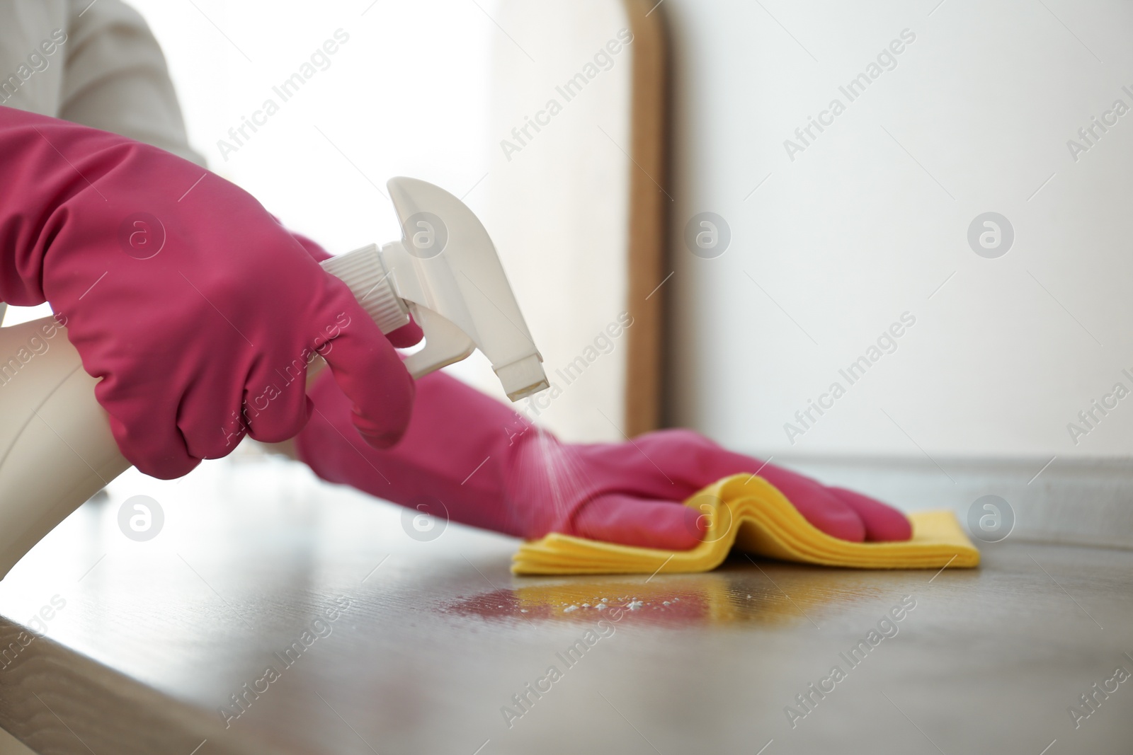 Photo of Woman in gloves cleaning wooden table with spray detergent and rag indoors, closeup