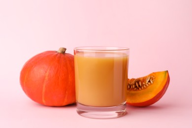 Photo of Tasty pumpkin juice in glass, whole and cut pumpkins on pale pink background