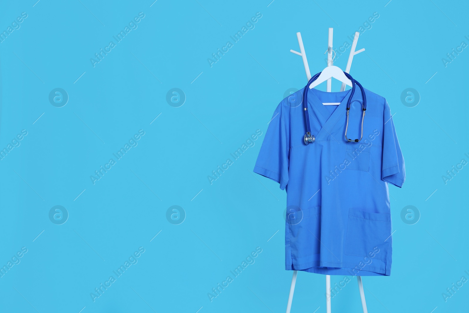Photo of Medical uniform and stethoscope hanging on rack against light blue background. Space for text