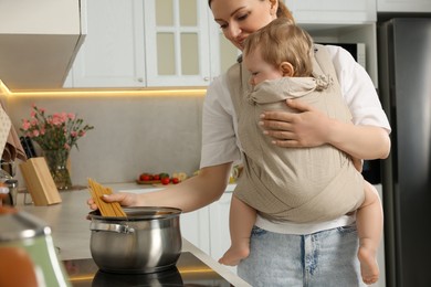 Mother holding her child in sling (baby carrier) while cooking pasta in kitchen