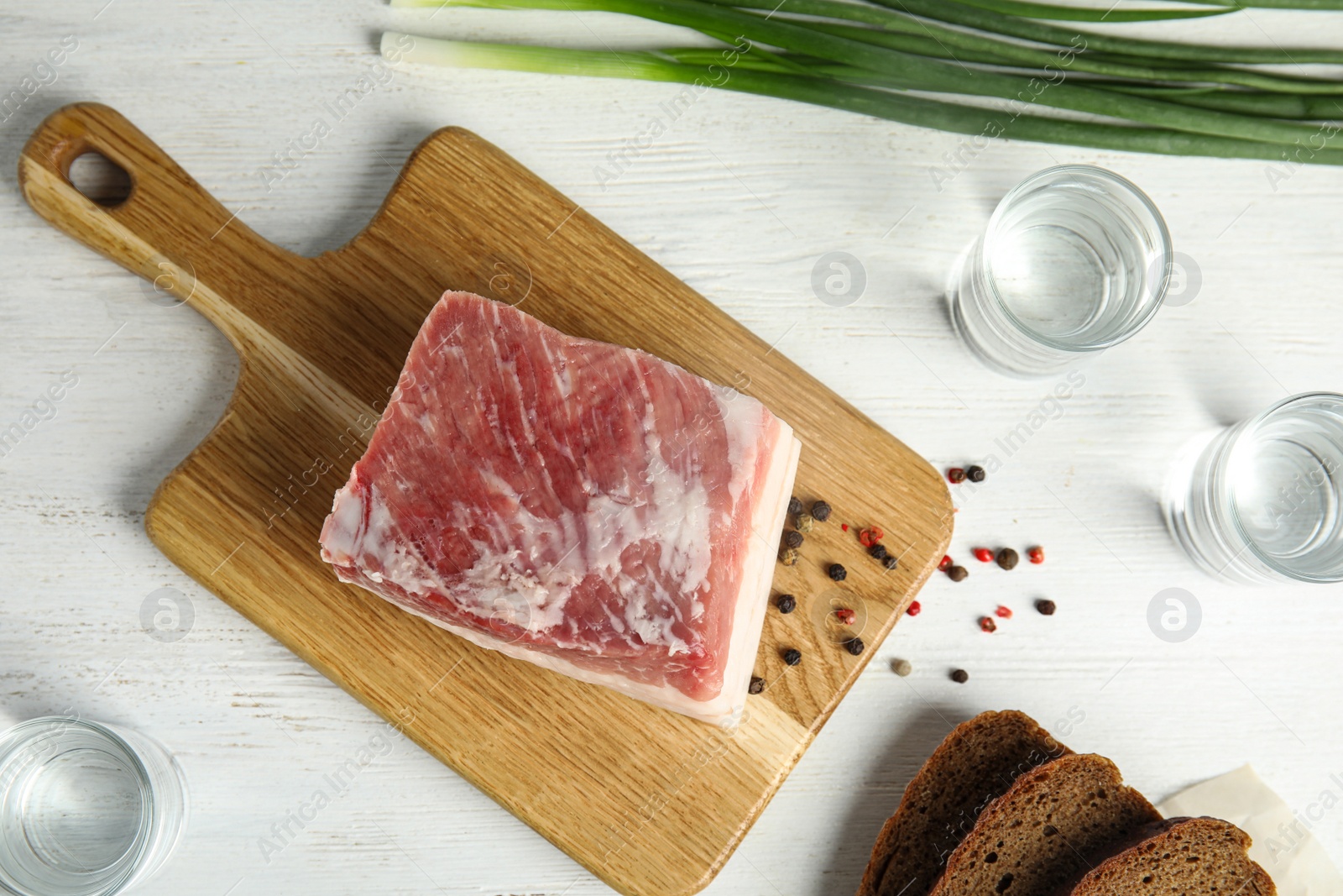 Photo of Board with pork fatback, onion, bread and shots of vodka on white wooden background, flat lay