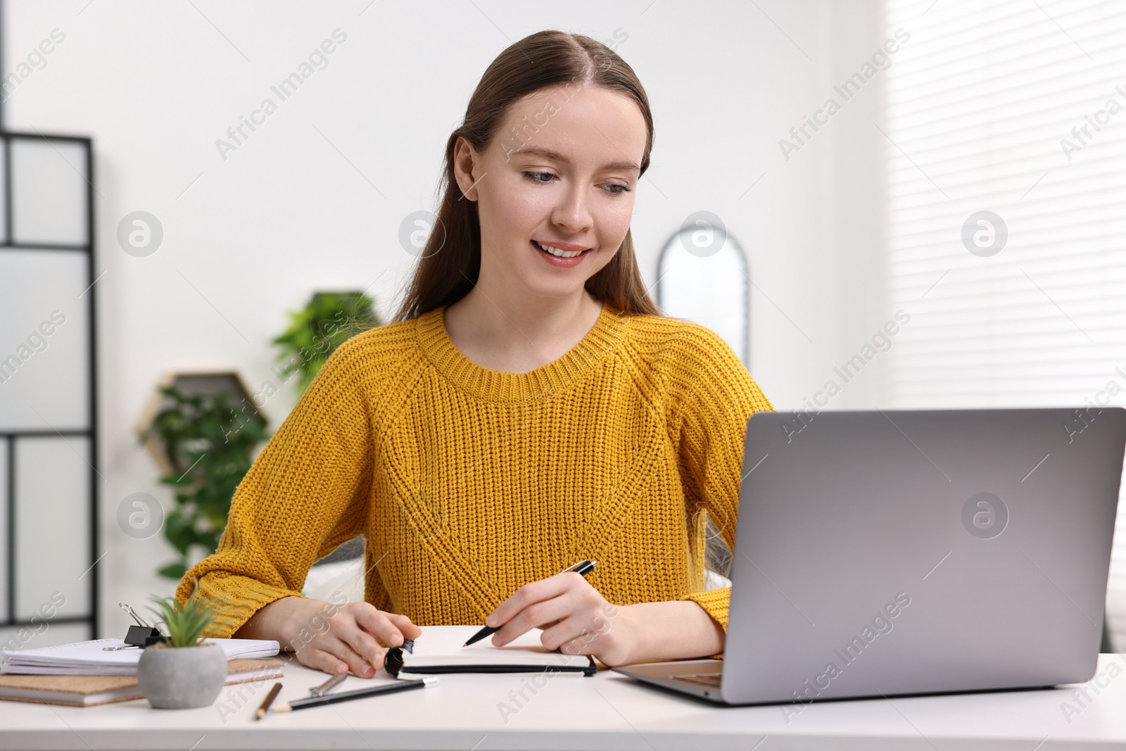 Photo of E-learning. Young woman taking notes during online lesson at white table indoors