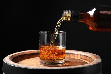 Photo of Pouring whiskey from bottle into glass on wooden barrel against black background, closeup