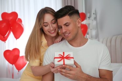 Woman presenting gift to her boyfriend in room decorated with heart shaped balloons. Valentine's day celebration