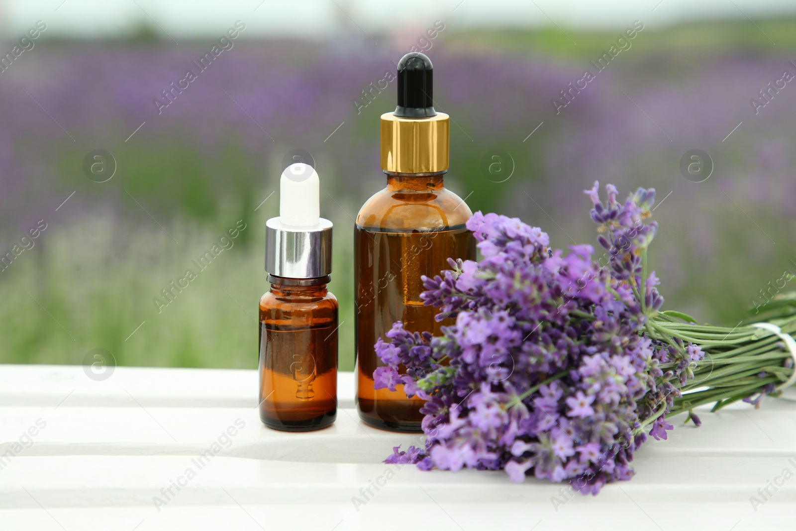 Photo of Bottles of essential oil and lavender flowers on white wooden table in field