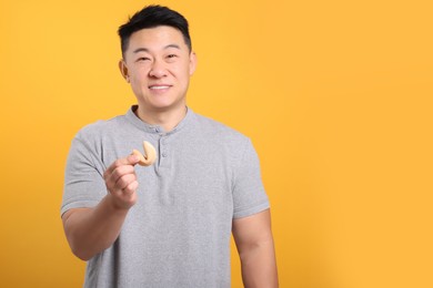 Photo of Asian man holding tasty fortune cookie with prediction on orange background. Space for text