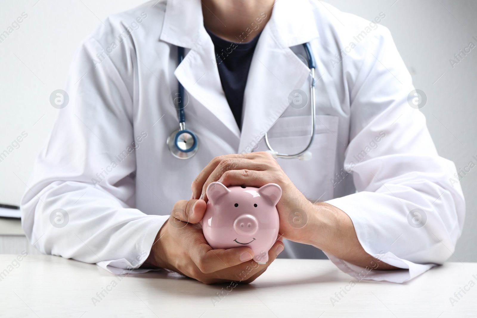 Photo of Doctor with piggybank at white table indoors, closeup. Medical insurance concept