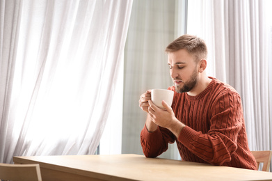 Young man with cup of hot drink near window at cafe