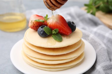 Pouring honey from dipper onto delicious pancakes with berries and mint on light grey table, closeup