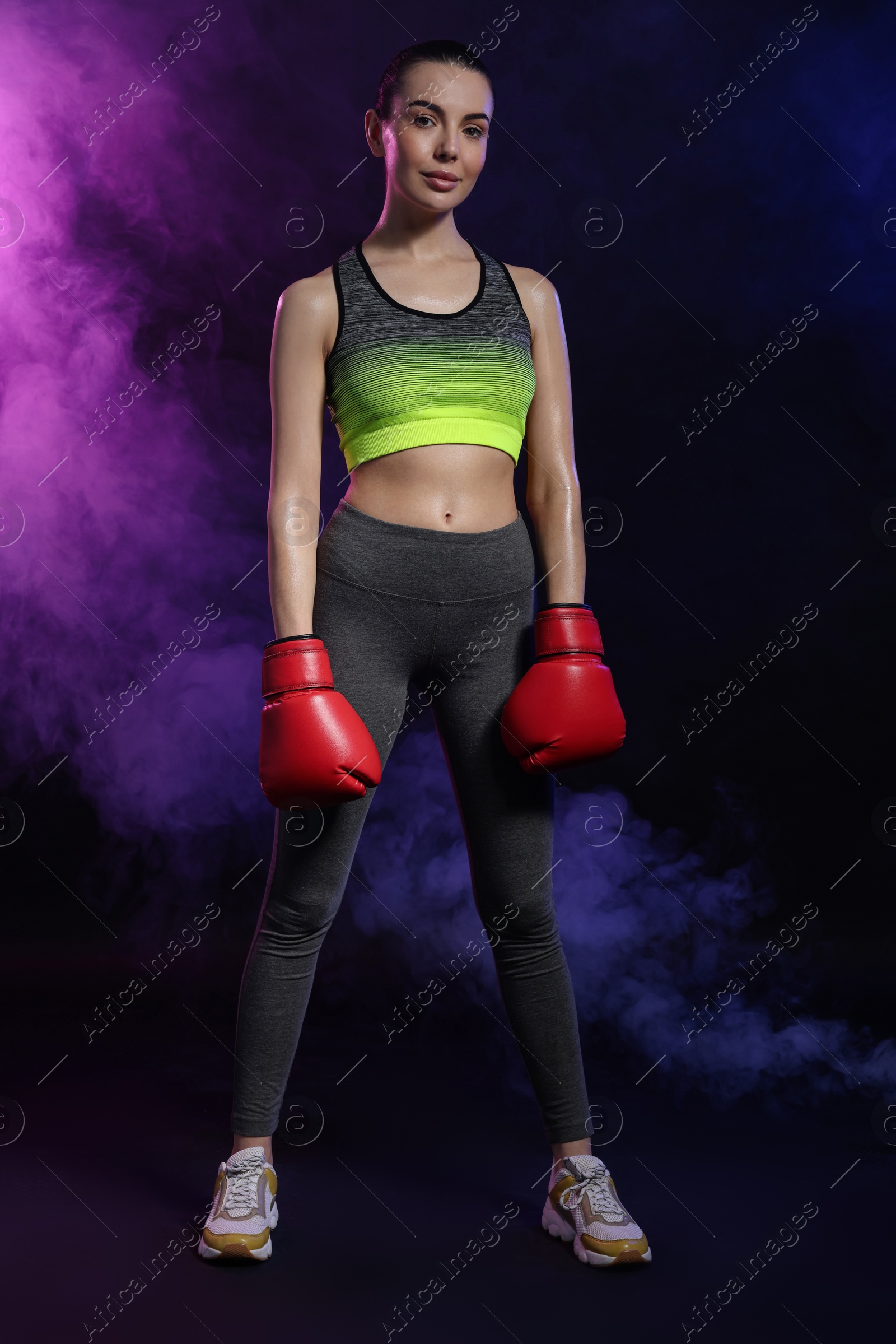 Photo of Beautiful woman wearing boxing gloves in color lights and smoke on black background