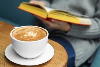 Woman reading book at table, focus on cup of coffee