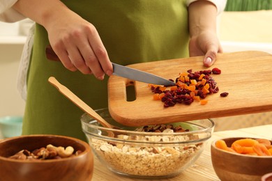 Photo of Making granola. Woman adding dried apricots and cherries into bowl with oat flakes at table in kitchen, closeup