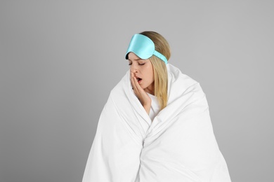 Photo of Young woman in sleeping mask wrapped with soft blanket yawning on grey background