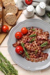 Photo of Slices of tasty fried bacon with rosemary and tomatoes served on wooden table, flat lay