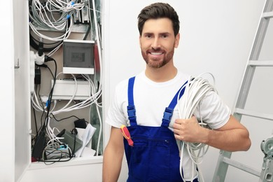 Smiling electrician with wires near open fuse box indoors