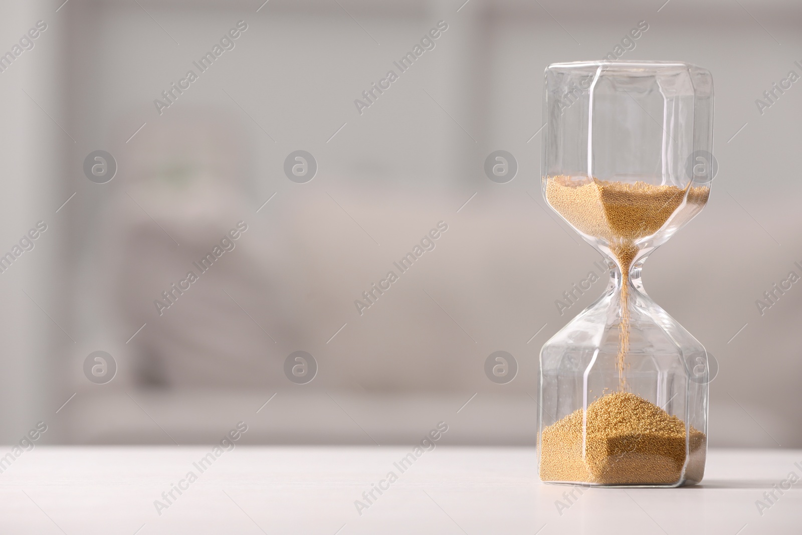 Photo of Hourglass with flowing sand on white table against blurred background. Space for text