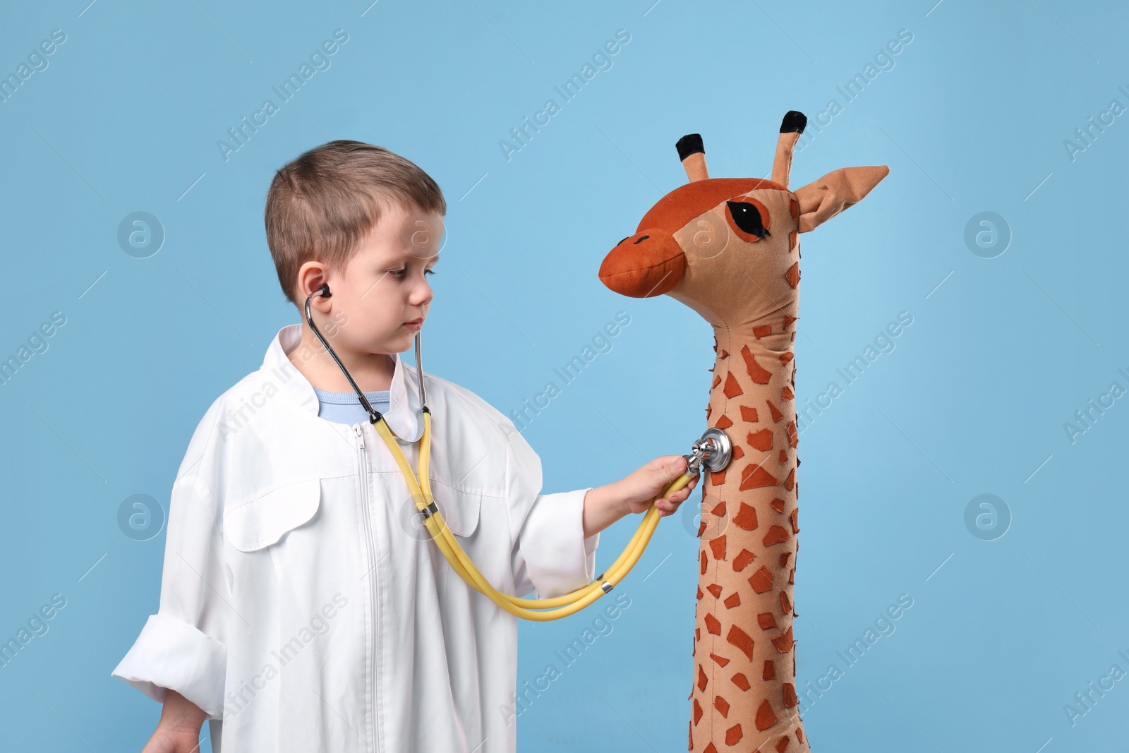 Photo of Cute little boy in pediatrician's uniform playing with stethoscope and toy giraffe on light blue background