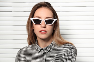 Young woman wearing stylish sunglasses against blinds