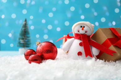Cute toy snowman, gift box and Christmas balls on snow against blurred background. Space for text