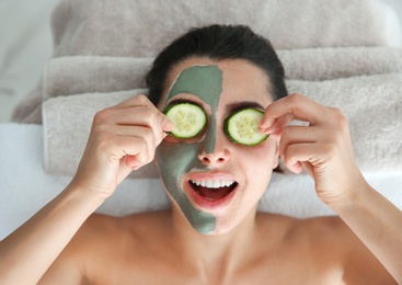 Pretty woman with clay mask on her face holding cucumber slices in spa salon, above view