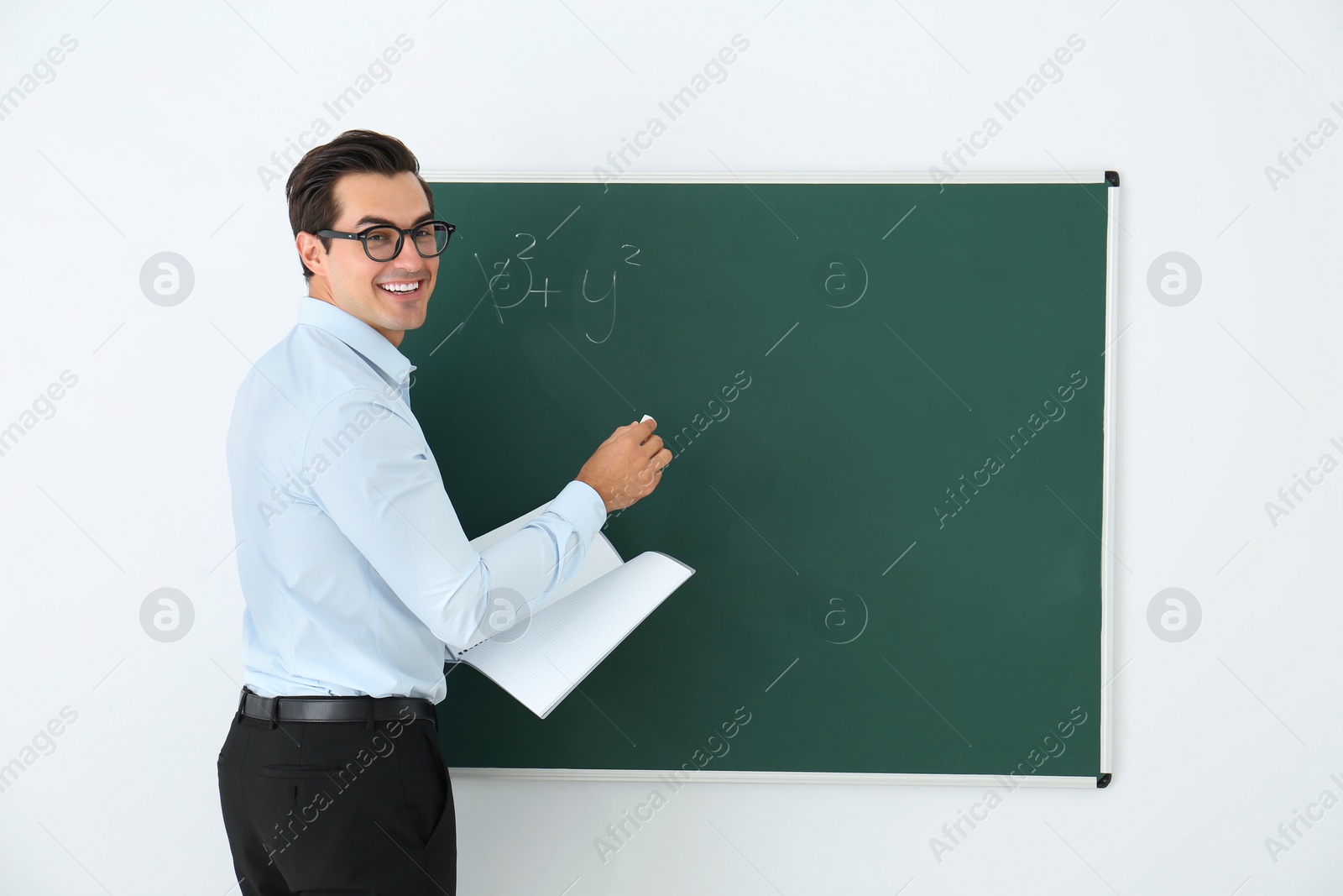 Photo of Young teacher with book near blank chalkboard in classroom. Space for text