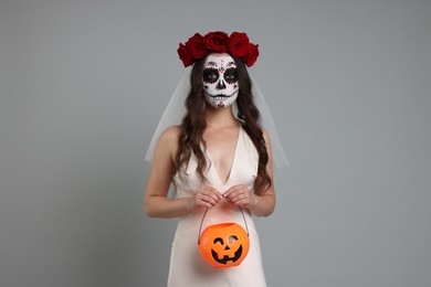 Photo of Young woman in scary bride costume with sugar skull makeup, flower crown and pumpkin bucket on light grey background. Halloween celebration
