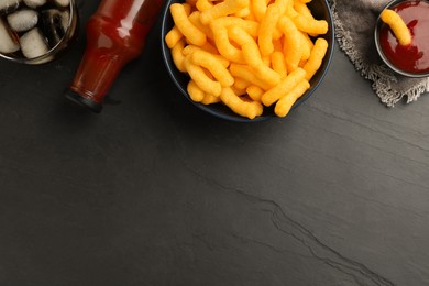 Bowl with crunchy cheesy corn snack, ketchup and refreshing drink on black table, flat lay. Space for text