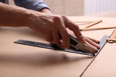 Photo of Man cutting chip board with utility knife and ruler indoors, closeup