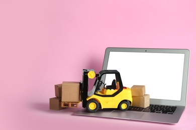 Laptop, forklift model and carton boxes on pink background. Courier service