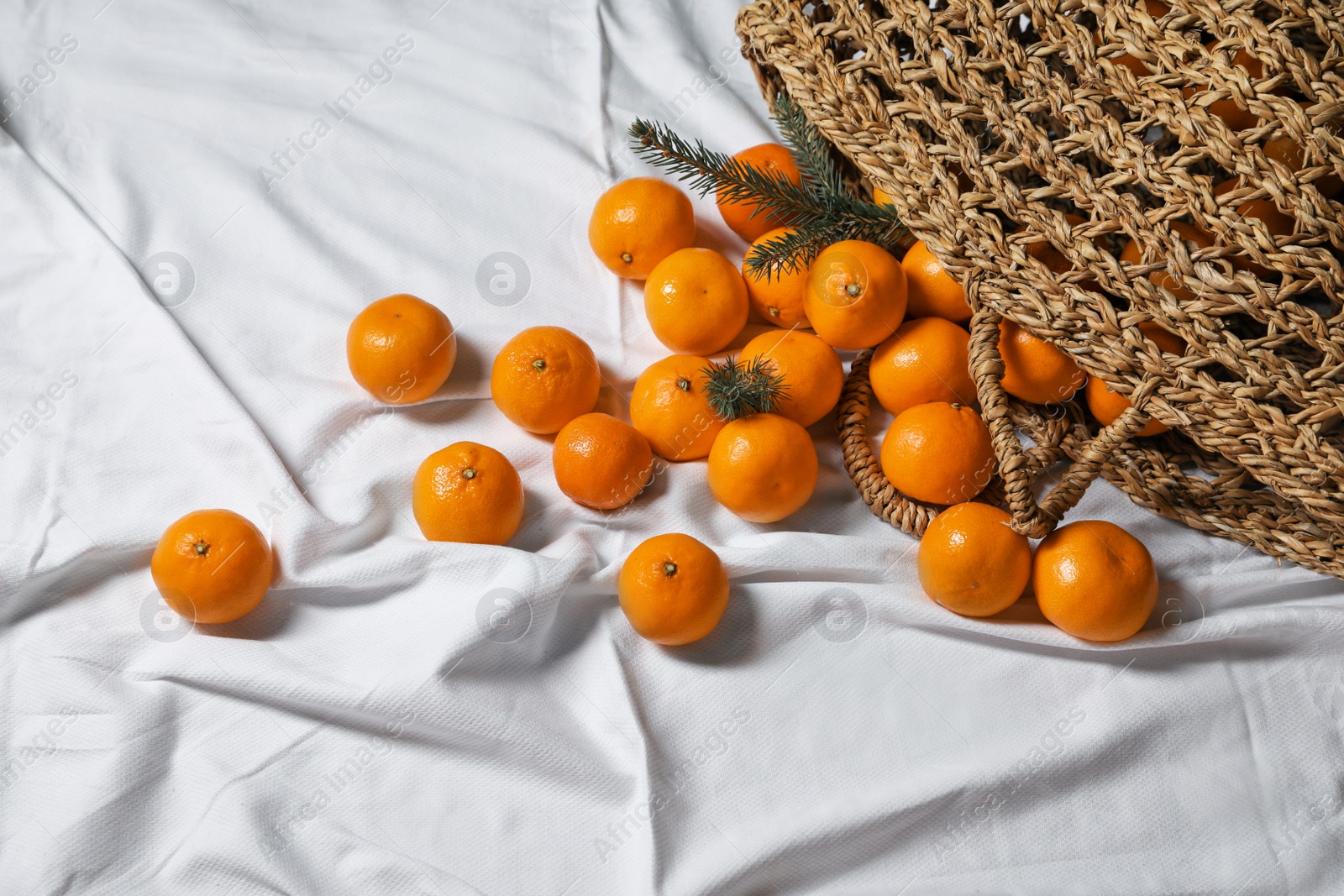 Photo of Stylish wicker bag with ripe tangerines on white bedsheet, above view