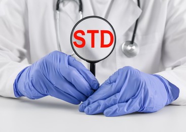 Doctor holding magnifying glass with abbreviation STD at white table, closeup