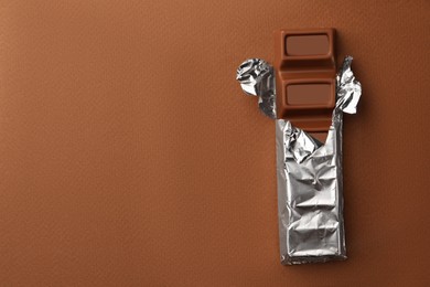 Delicious chocolate bar wrapped in foil on brown background, top view. Space for text