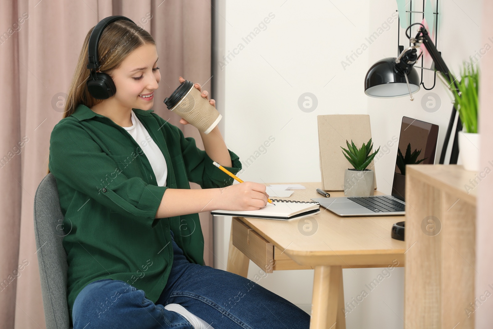 Photo of Teenage girl with headphones and cup of drink writing in notebook at wooden table indoors