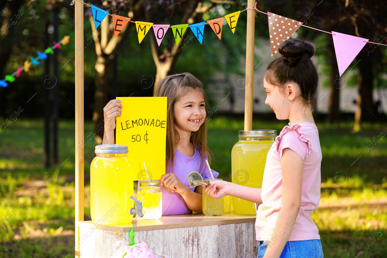 Photo of Cute little girl selling natural lemonade to kid in park. Summer refreshing drink