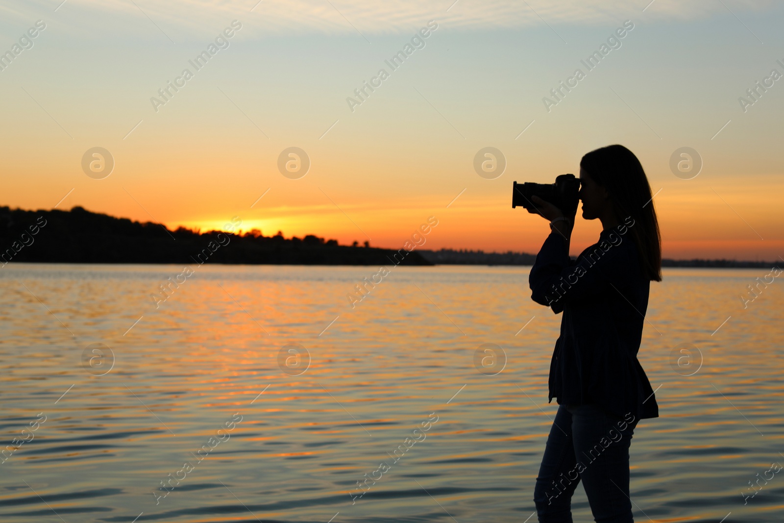 Photo of Young female photographer taking photo of riverside sunset with professional camera outdoors