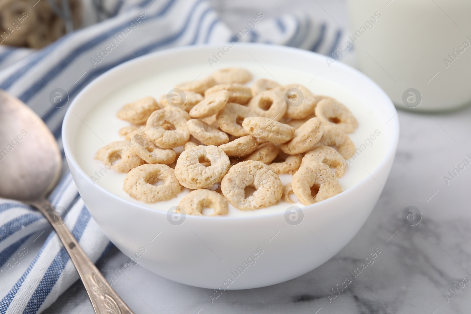 Photo of Breakfast cereal. Tasty corn rings with milk in bowl and spoon on white marble table, closeup