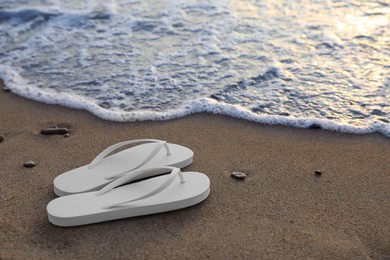 Photo of Stylish white flip flops on sand near sea. Space for text