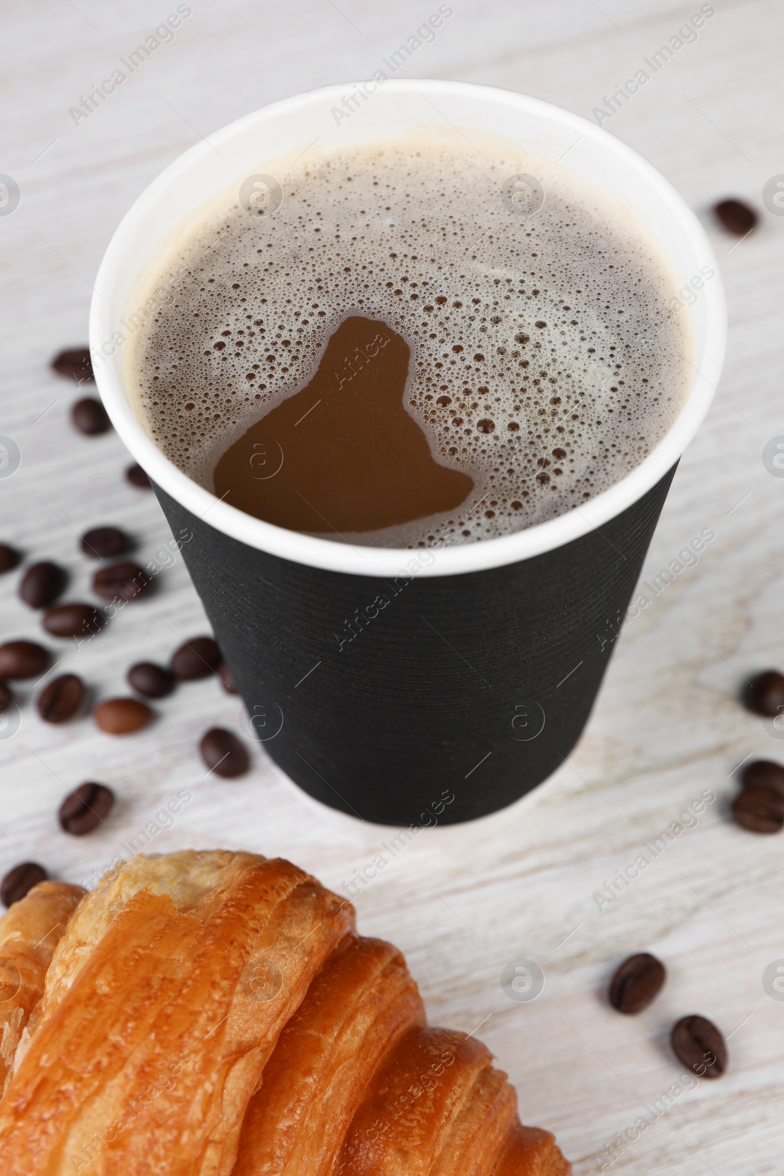 Photo of Coffee to go. Paper cup with tasty drink, croissant and beans on white wooden table, closeup