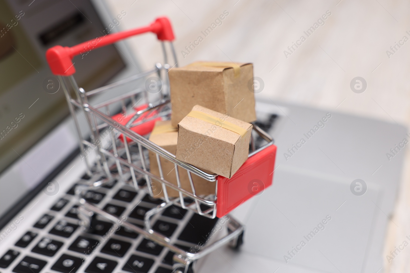 Photo of Internet store. Small cardboard boxes, shopping cart and laptop on light wooden table, closeup. Space for text