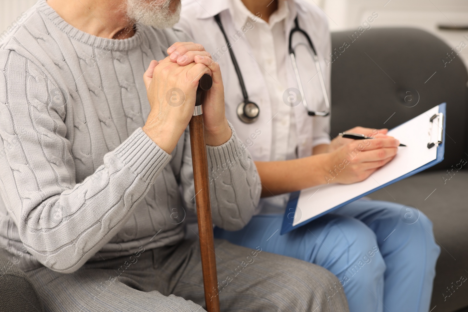 Photo of Nurse with clipboard assisting elderly patient on sofa indoors, closeup