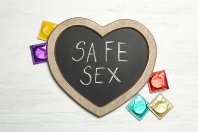 Photo of Colorful condoms and heart shaped blackboard with words SAFE SEX on white wooden background, flat lay