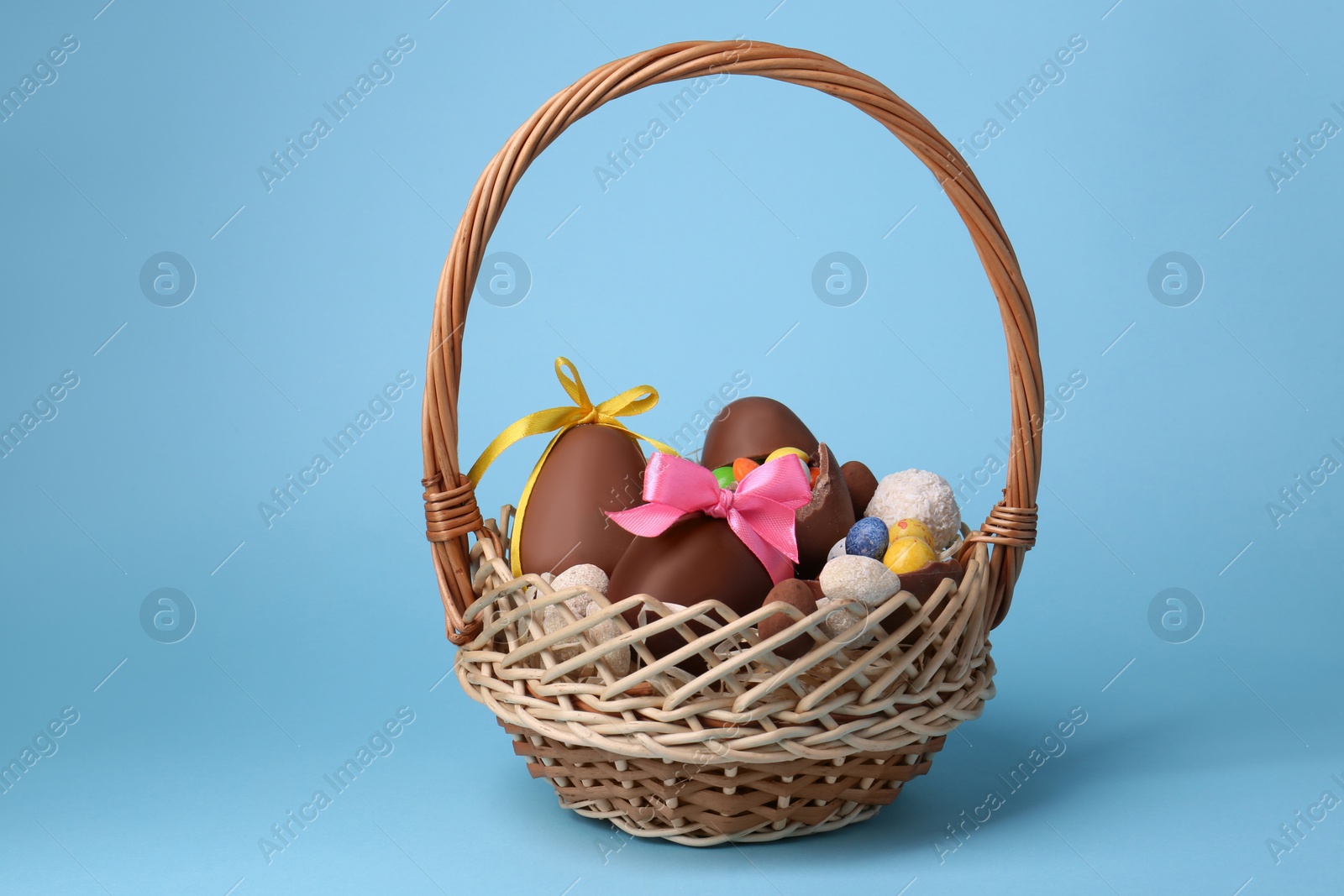 Photo of Wicker basket with tasty chocolate Easter eggs and different candies on light blue background