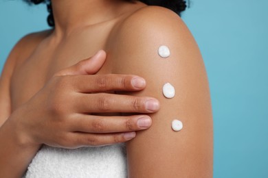 Photo of Young woman applying body cream onto shoulder on light blue background, closeup