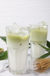 Photo of Glasses of tasty iced matcha latte, bamboo whisk and leaves on white marble table